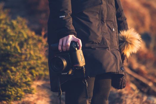 Side view of crop anonymous woman in outerwear holding professional contemporary photo camera while standing with knitted hat with pompon near green bush in daylight