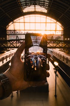 From above of crop anonymous male taking photo of railroad with trains on cellphone while standing in modern building in front of window in sunshine