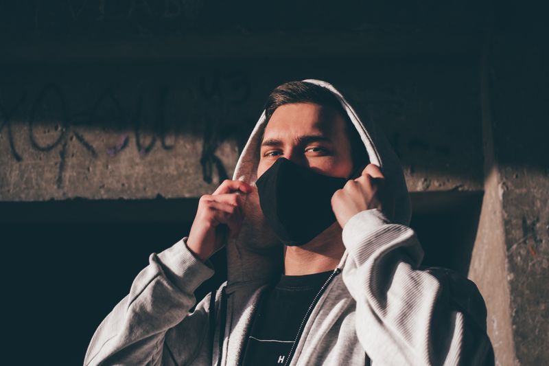 Young unrecognizable trendy man in mask and hoodie looking at camera while standing near rough concrete wall.