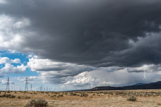 Breathtaking view of thick dark clouds on sky with lightning above uneven field with growing plants near metal towers and mountains in gloomy weather