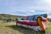 Old unnecessary dirty couch on grass meadow near hill and growing plants under cloudy sky in summertime in daylight in countryside