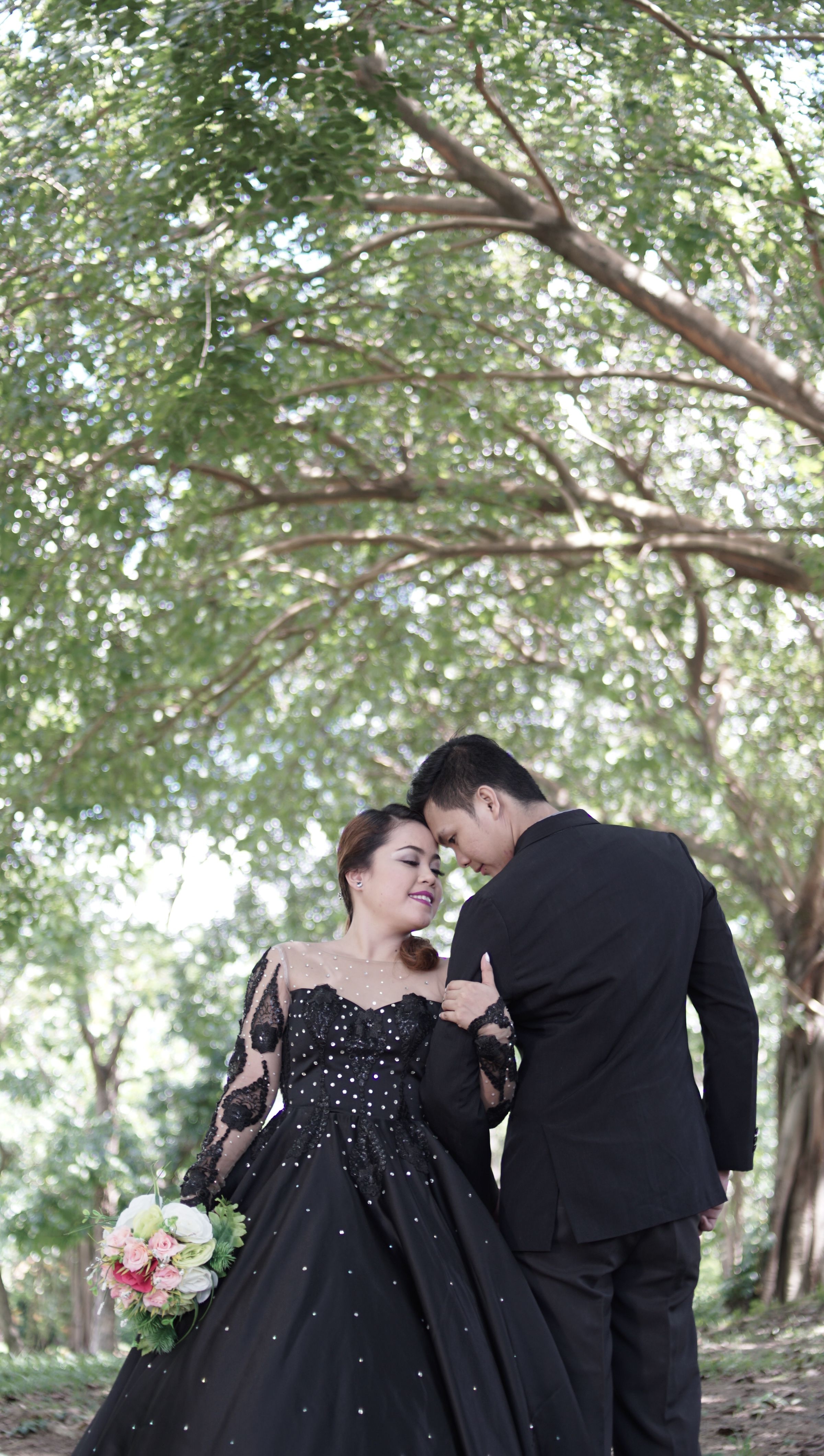 A couple in black wedding attire enjoy a romantic moment outdoors, under lush trees.