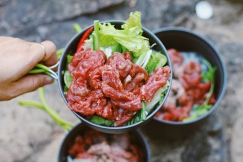 From above of crop unrecognizable person showing small pan with fresh green salad and sliced raw beef