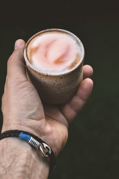 From above crop anonymous male with bracelet holding small cup of cappuccino with foam in hand