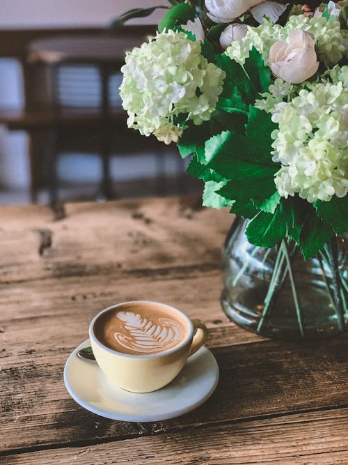 Free From above cup of hot cappuccino with latte art placed on wooden table and arranged with gentle bouquet of fresh hydrangea flowers Stock Photo