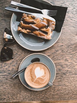Top view sunglasses and cup of coffee with heart shaped latte art served on wooden table in cafe with plate of delicious sweet pie topped with chocolate syrup