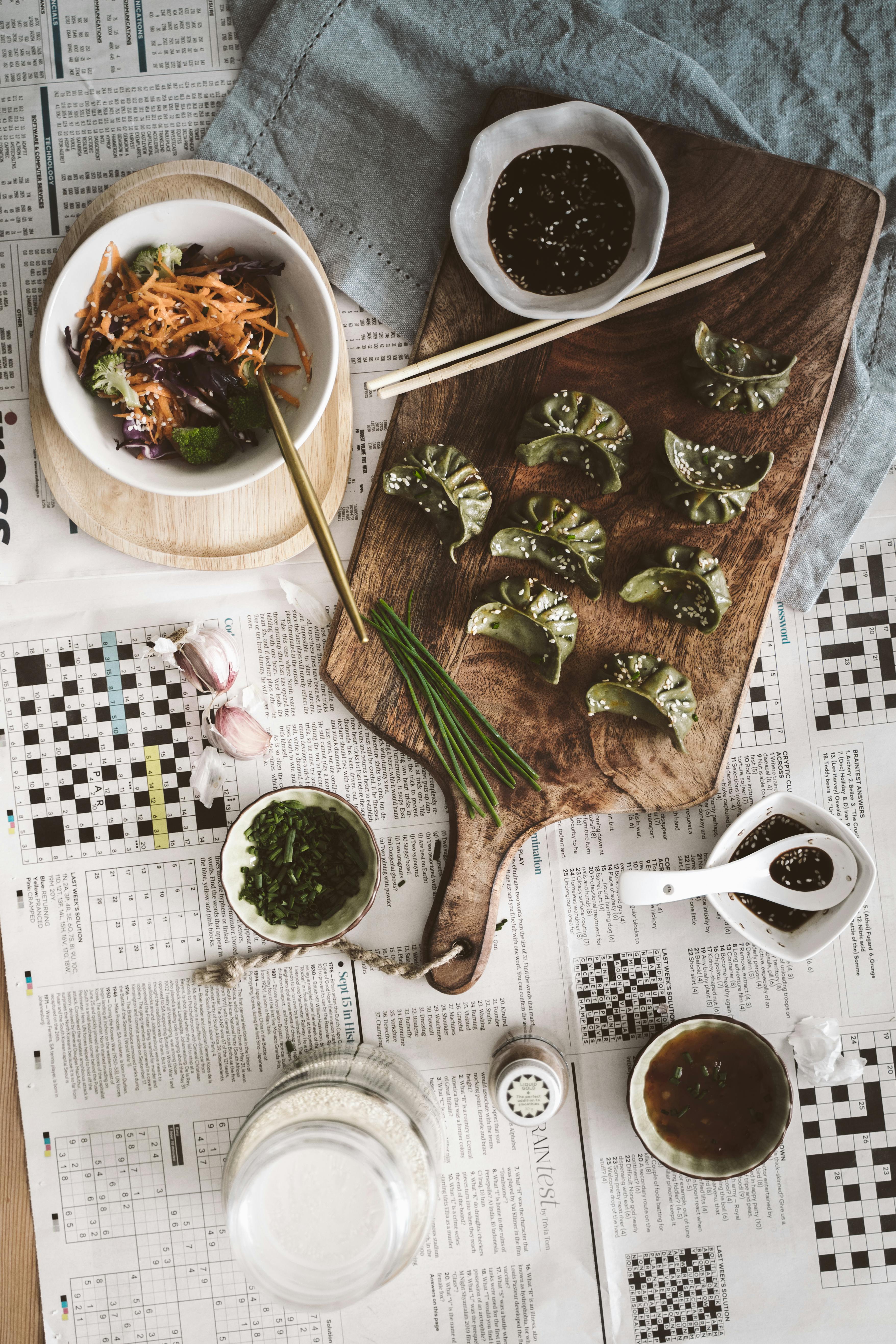 Flatlay of Asian dumplings with sauce dips and salad on wooden board and newspapers.