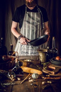 Crop anonymous bearded male chef in apron with pan in hand and standing at table with various natural ingredients and kitchenware