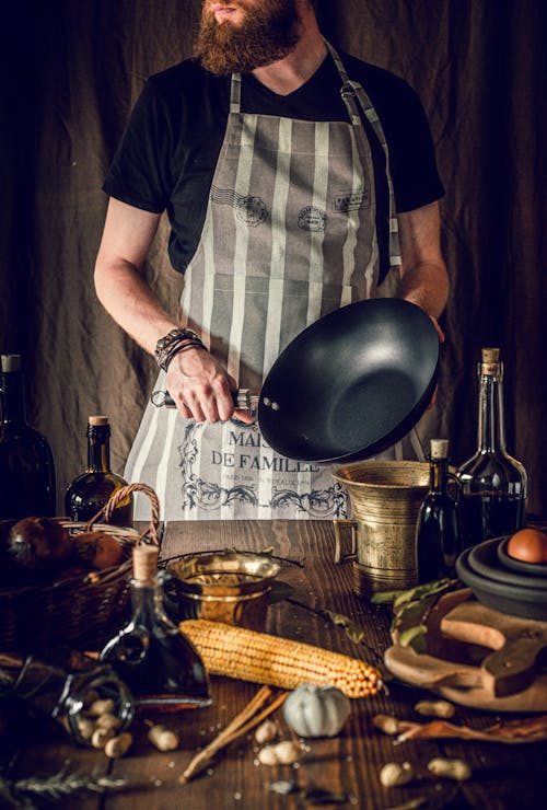Crop unrecognizable bearded male cook in apron holding frying pan in hands while preparing healthy delicious dish in rustic kitchen