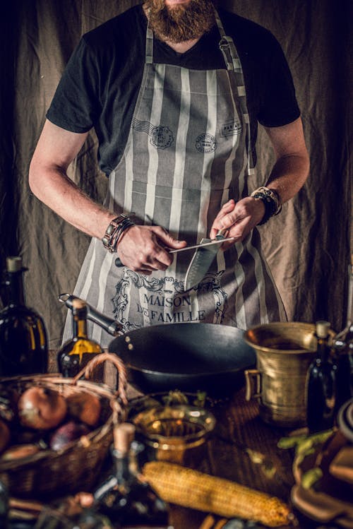 Crop young bearded male chef in apron sharpening knife while preparing delicious traditional dish with onions and corn in kitchen