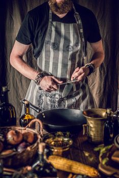 Crop young bearded male chef in apron sharpening knife while preparing delicious traditional dish with onions and corn in kitchen