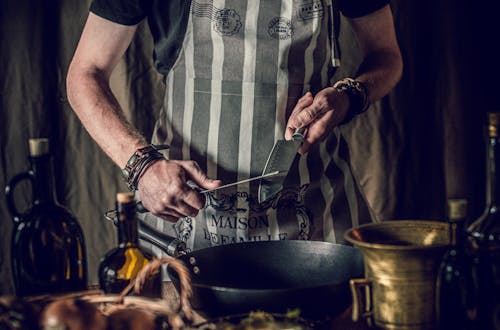 Crop anonymous male cook in apron holding sharp knives in hands while standing at table with frying pan and oil bottles and preparing lunch