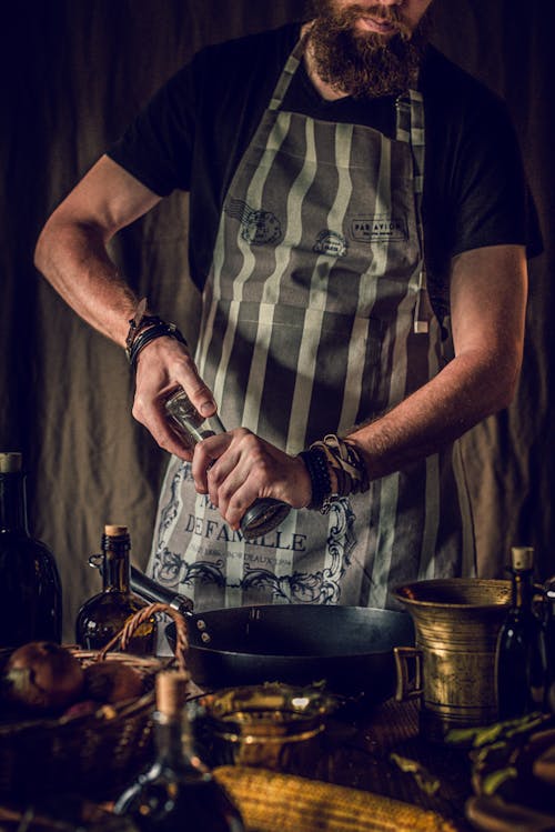 Crop anonymous serious bearded male chef adding pepper using mill in frying pan while preparing seasonal dish in kitchen