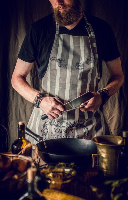 Free Crop anonymous young male cook in apron standing at table with pan and various products and sharpening knife before preparing healthy dinner in restaurant kitchen Stock Photo