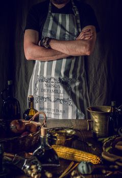 Crop faceless male in apron standing with crossed arms near table with assorted fresh groceries and vintage kitchenware
