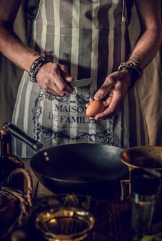 A man in an apron cracks an egg into a pan in a rustic kitchen setting.