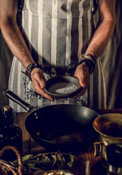 Crop man in striped apron holding plate of condiment while cooking in frying pan in bright room
