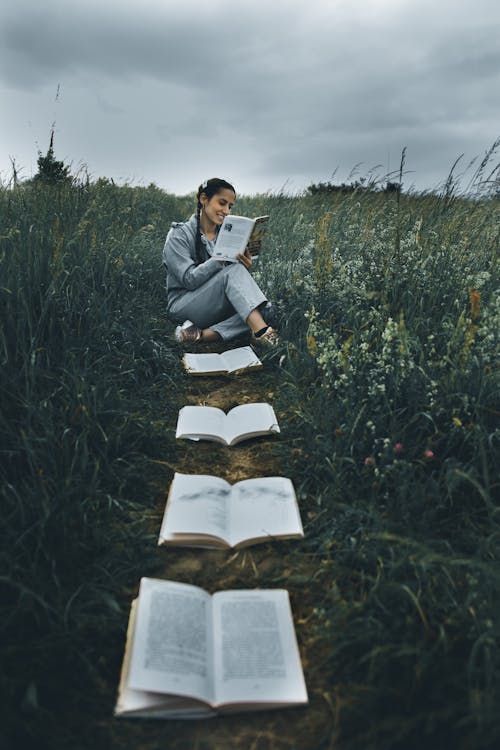 From above of cheerful ethnic woman reading textbook while sitting with crossed legs on path with different open books between grass under cloudy sky