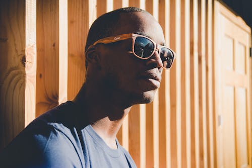 Pria Bersandar Di Dinding Kayu Coklat Sambil Mengenakan Kacamata Hitam Dan Menghadapi Sinar Matahari