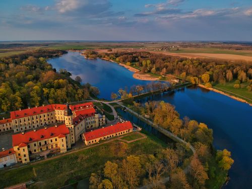 Free Picturesque drone view of ancient Nesvizh Castle with red roofs located on shore of blue river surrounded by lush colorful autumn trees at sunset in Belarus Stock Photo