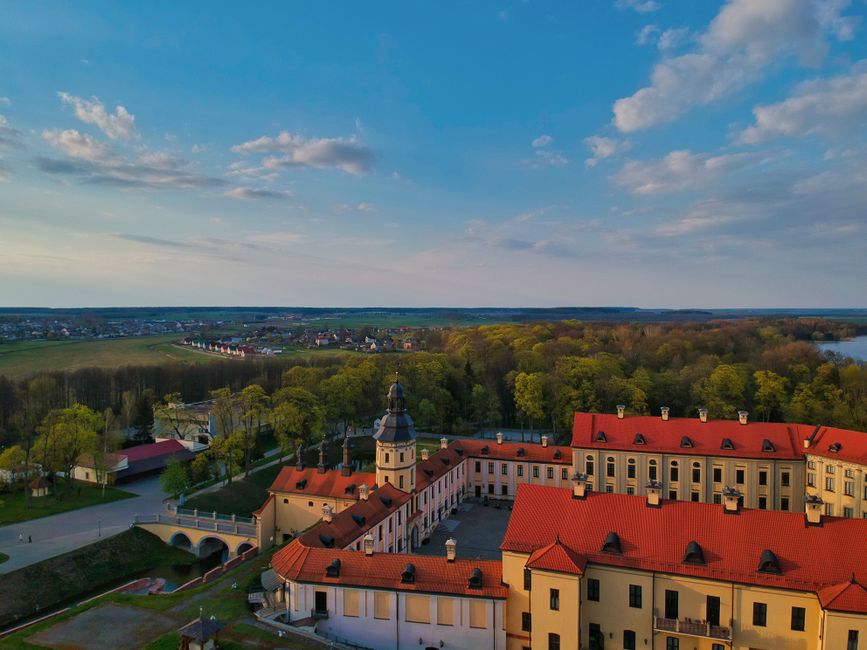 Drone view of red roofed historic Nesvizh Castle surrounded by lush autumn trees against cloudy blue sky