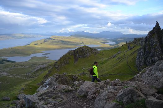 Modern backpacker walking on rocky slope of mountain range with lakes and highlands on background