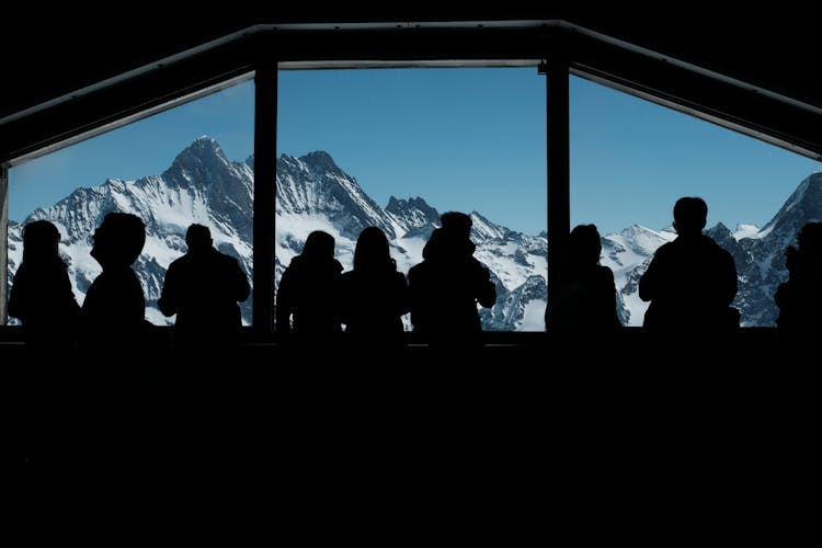 A Silhouette Of People Standing Near The Glass Window With Mountain View