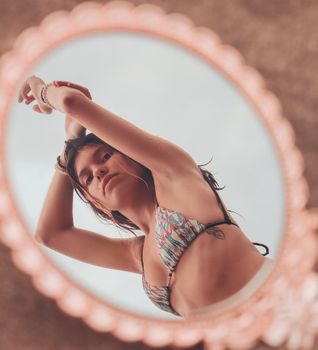 From above of slender female in swimsuit and wristbands standing with raised arms reflecting in mirror while looking at camera during summer vacation
