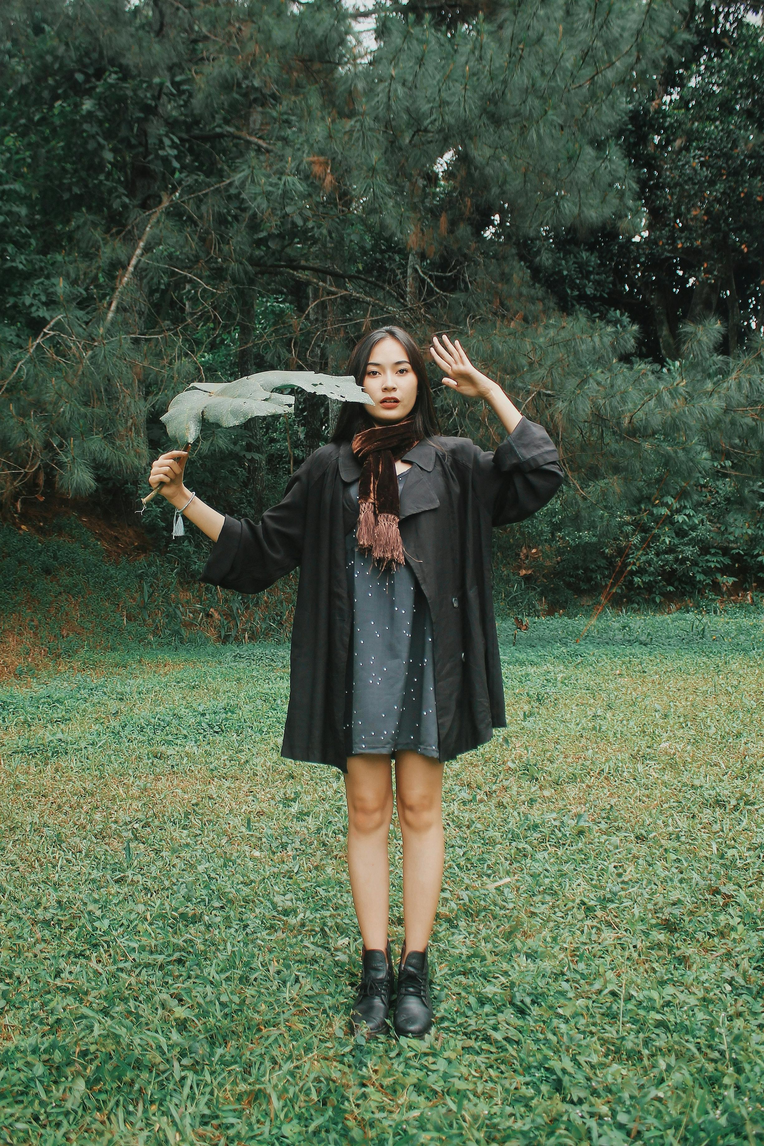Serious young ethnic woman saluting while holding tree leaf in city park and standing on grass looking at camera