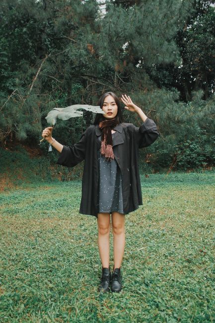 Serious young ethnic woman saluting while holding tree leaf in city park and standing on grass looking at camera