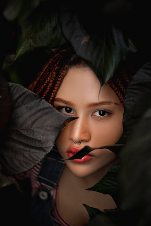 Sensual young female with braids and bright makeup standing near plant leaves in dark place and looking at camera
