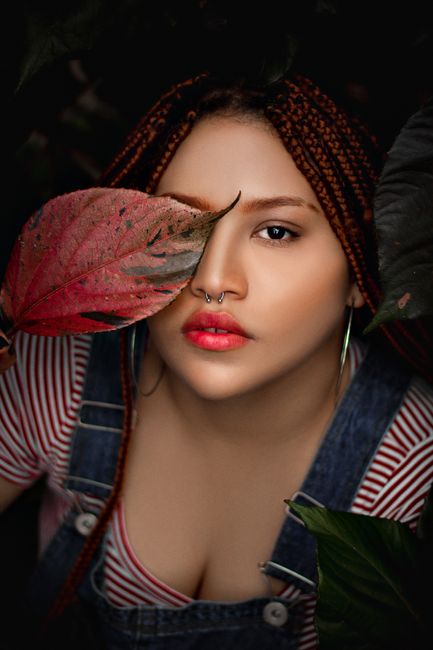 Alluring young female with bright makeup covering eye with leaf standing in dark place and looking at camera