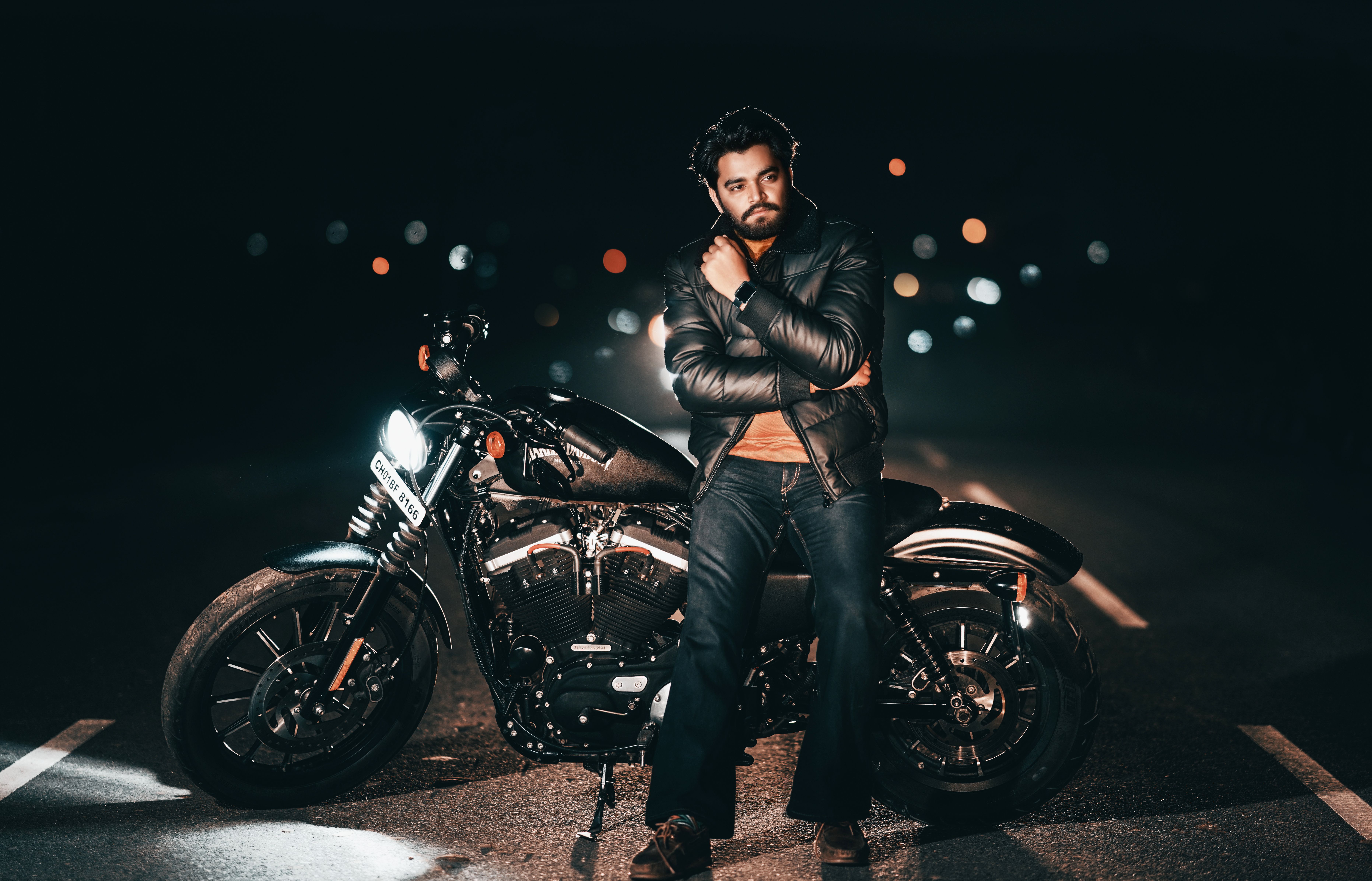 A man in a leather jacket poses with a motorcycle on a street at night, with bokeh lights in the background.