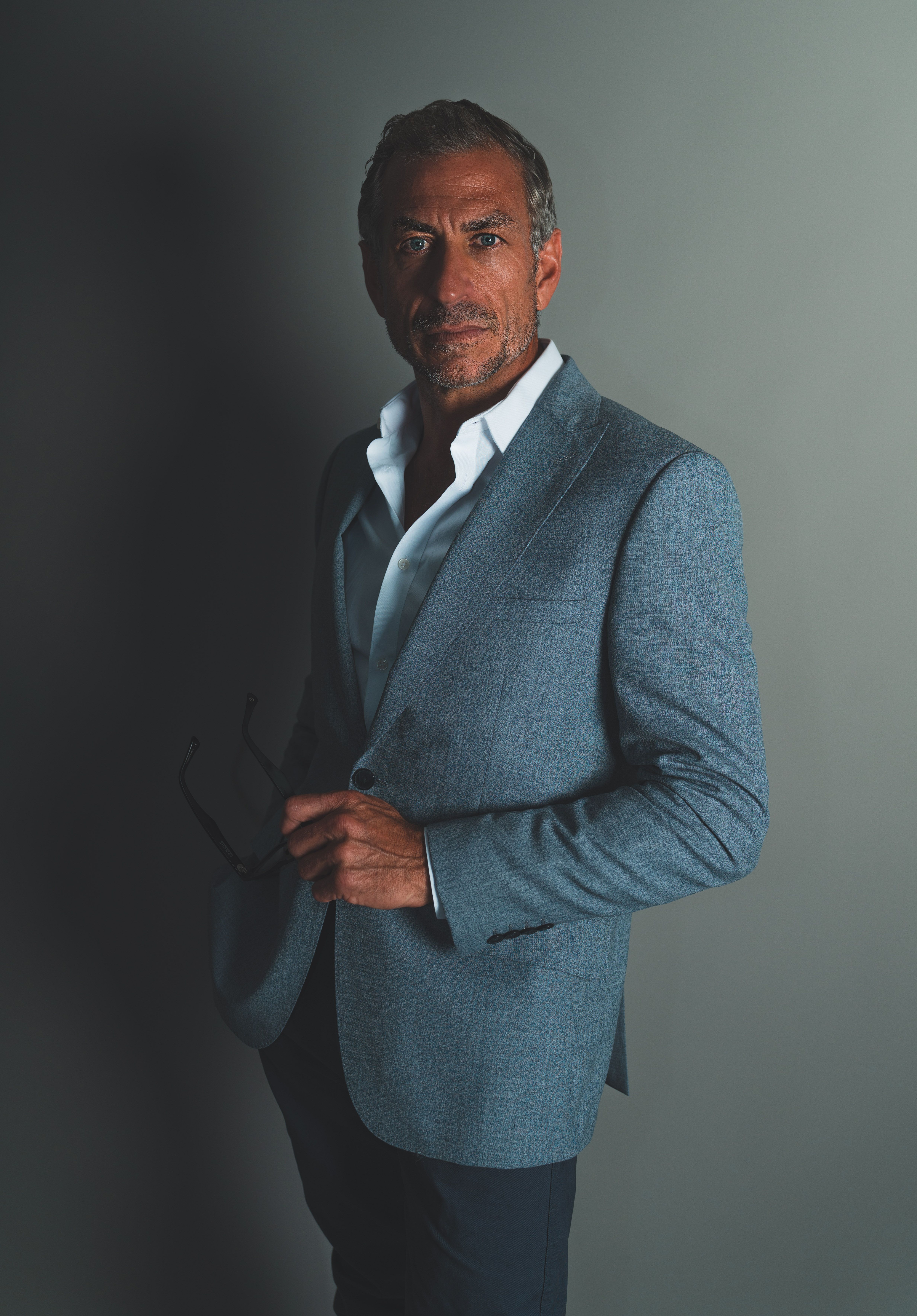Sophisticated adult man in a suit holding sunglasses against a studio background.