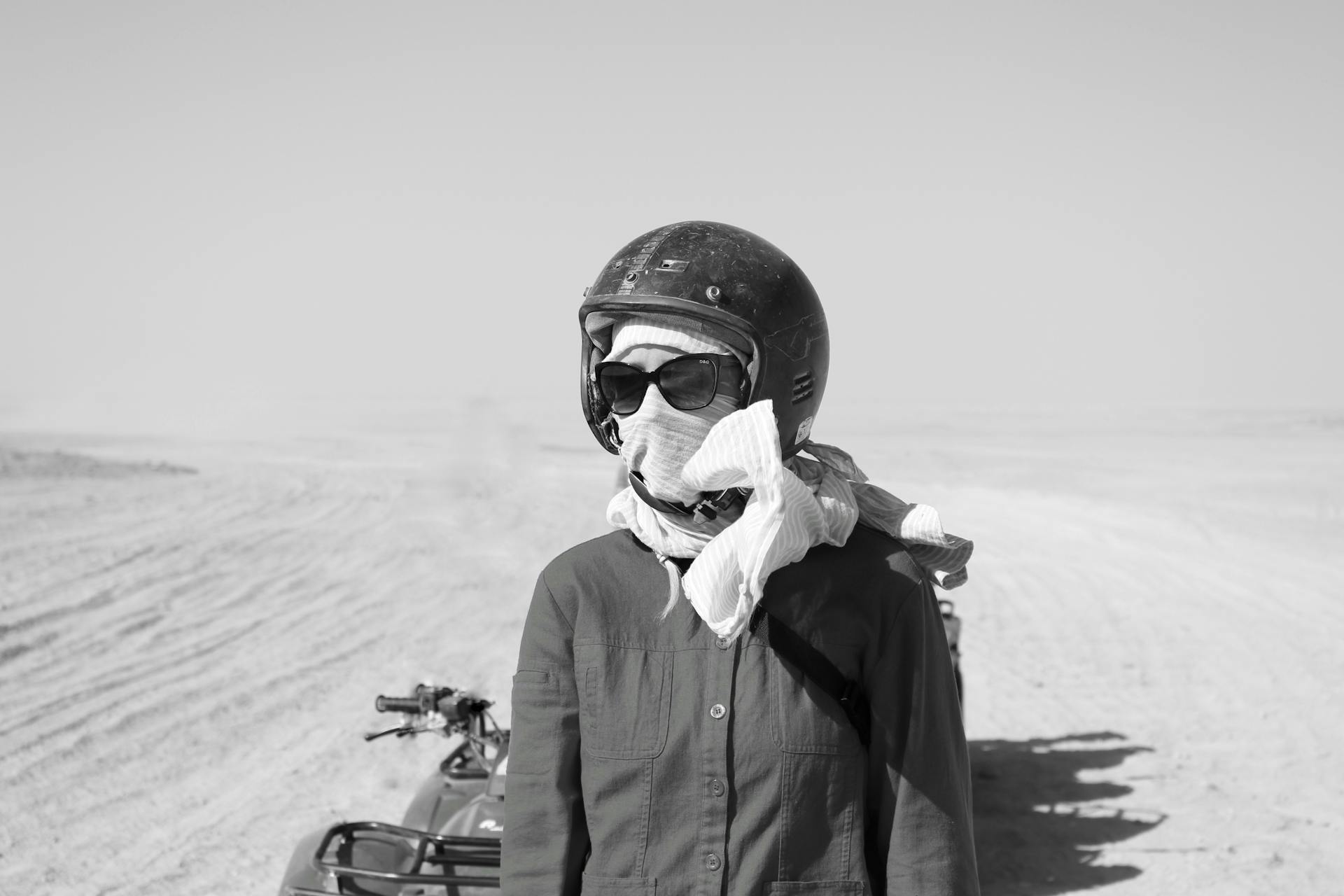 Black and white anonymous biker in helmet covering face with scarf and sunglasses standing near motorbike on sandy terrain