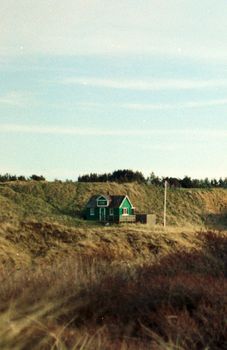 Small distant house with green walls located on grassy tranquil valley on hillside against blue sky