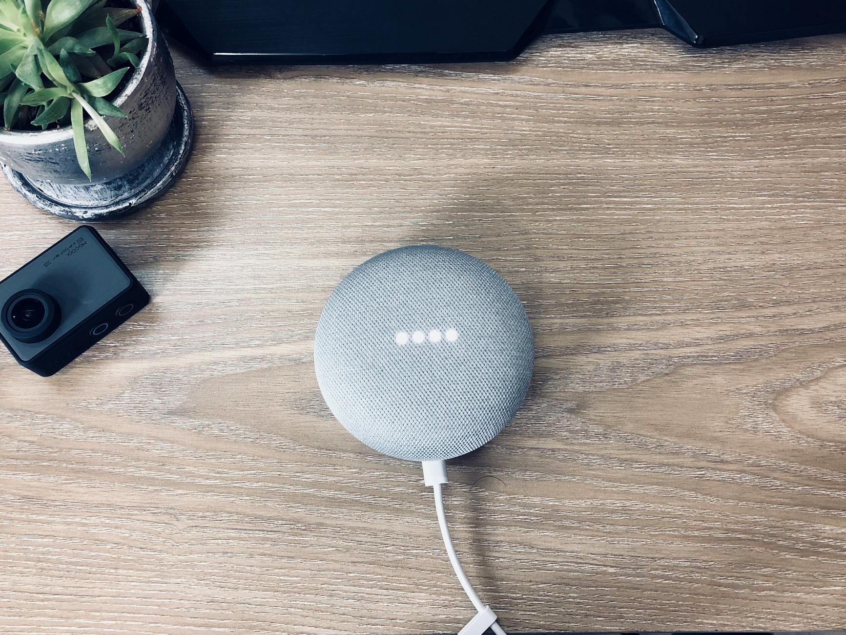 Top view of stylish smart speaker with connected white wire place on wooden table near small action camera and potted plant