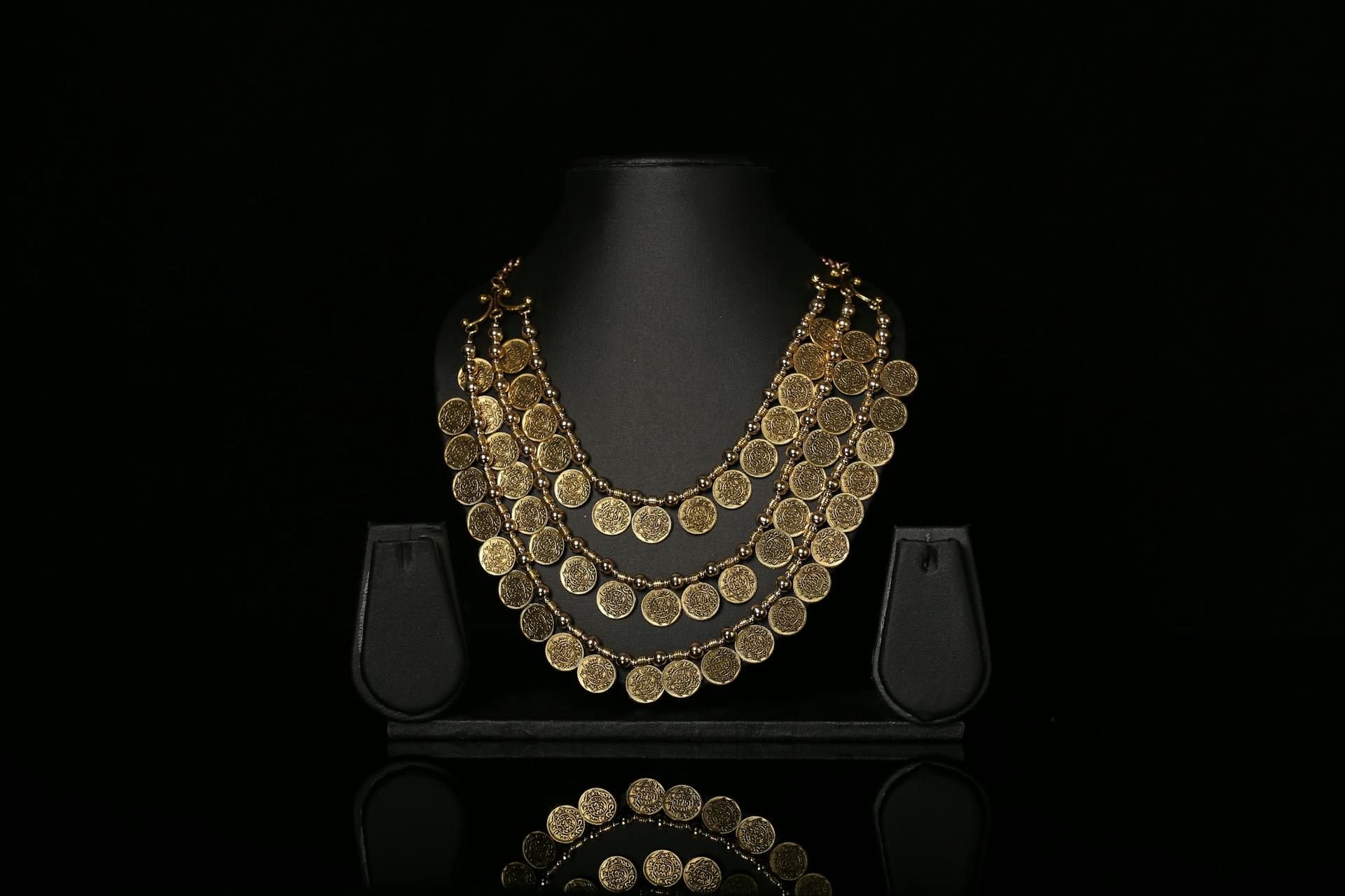 Luxurious gold coin necklace elegantly displayed on a black stand against a dark background.