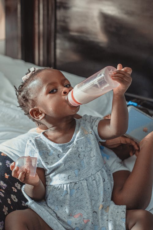 From above of cute little African American girl drinking milk from bottle and looking at camera while resting on bed