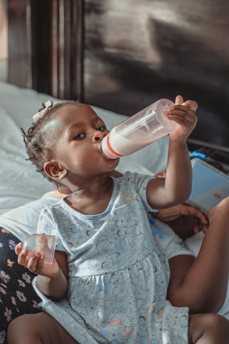 Adorable Black Baby Sitting On Bed And Drinking Milk From Bottle