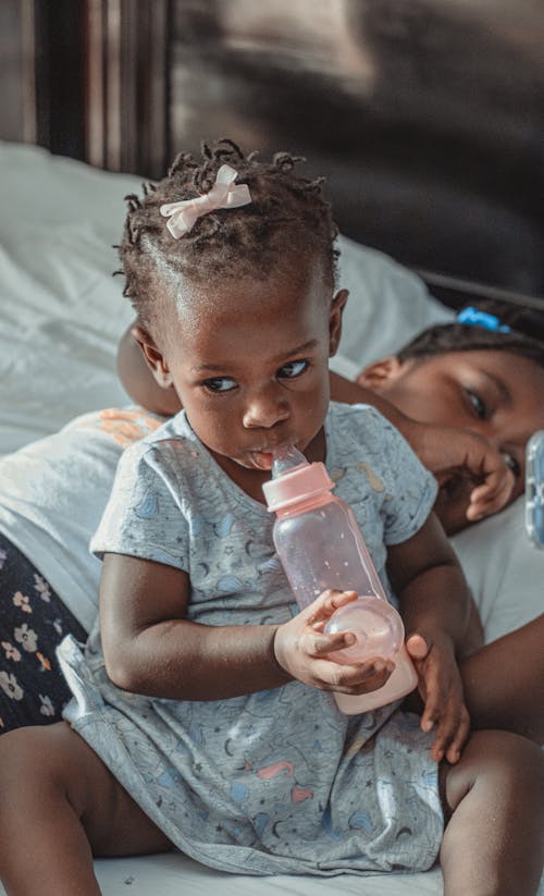 Cute African American child drinking milk from bottle on bed