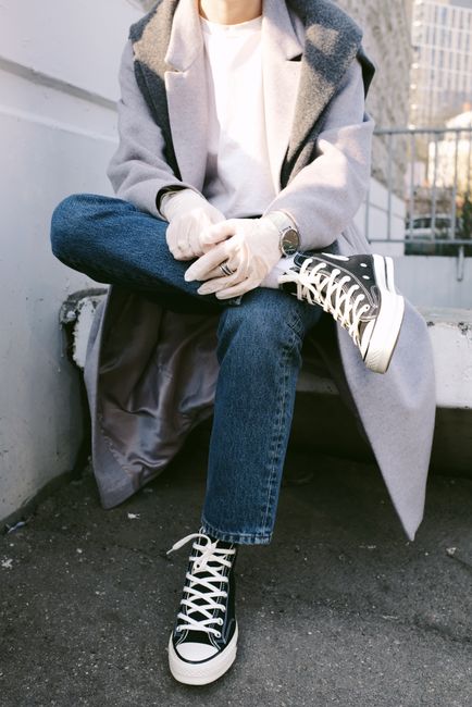 Person sitting outdoors wearing stylish sneakers and latex gloves, a gray coat, and jeans.