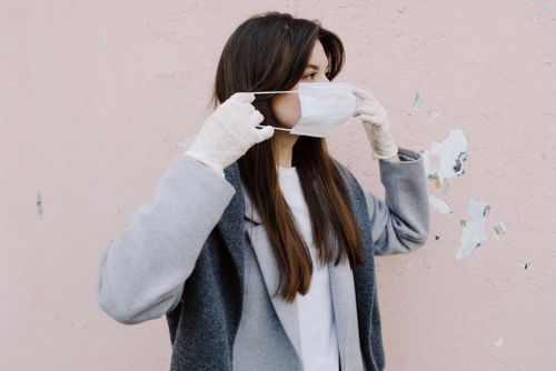 Free Young woman wearing a face mask and gloves for safety during the pandemic. Side view against a pink wall. Stock Photo