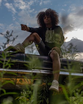 From below of young trendy African American male with Afro hair in stylish casual outfit sitting on car hood in meadow and looking at camera