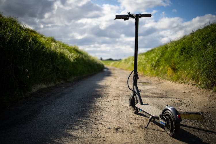 Electric Scooter On Rural Road In Grassland Against Cloudy Blue Sky