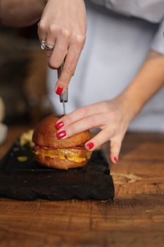 Close-up of a chef slicing a gourmet burger with vibrant nail polish indoors.