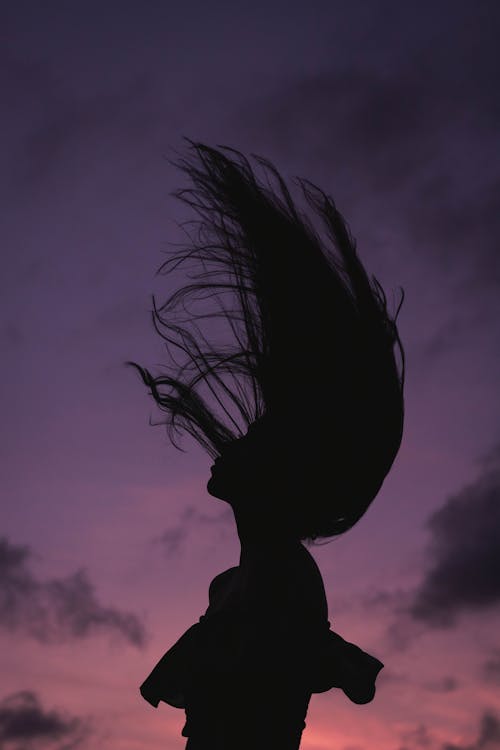 From below of unrecognizable female whipping hair during dark sundown sky in evening