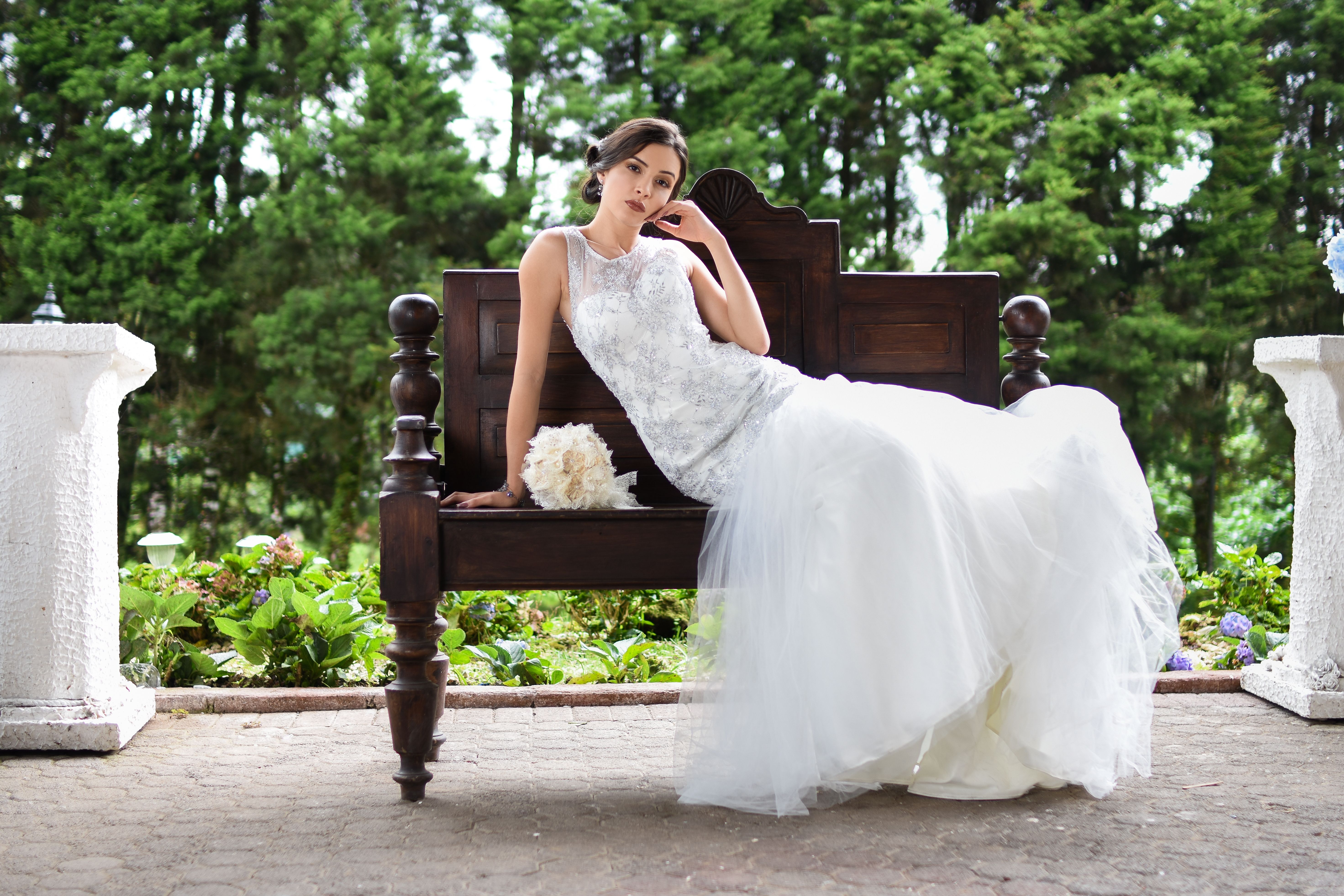 A beautiful bride in a white gown poses on a wooden bench in a lush garden setting.