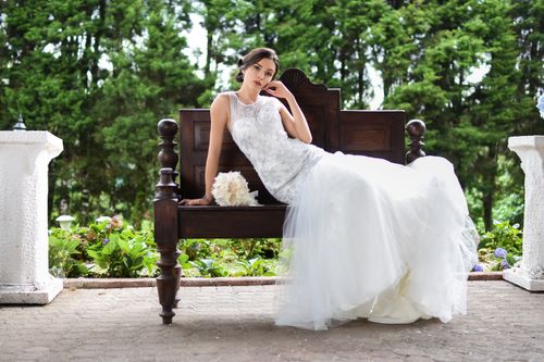 Free A beautiful bride in a white gown poses on a wooden bench in a lush garden setting. Stock Photo