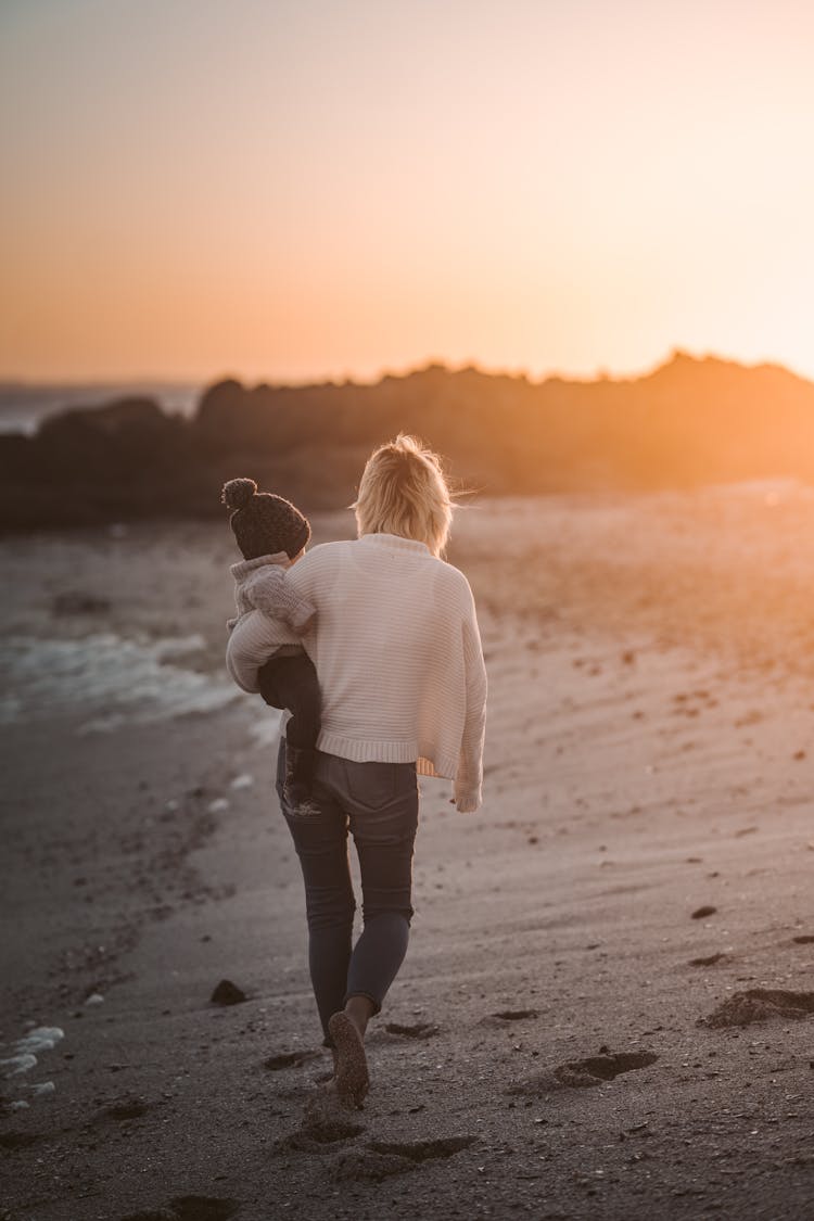 Back View Of A Mother Carrying Her Child While Walking On The Beach